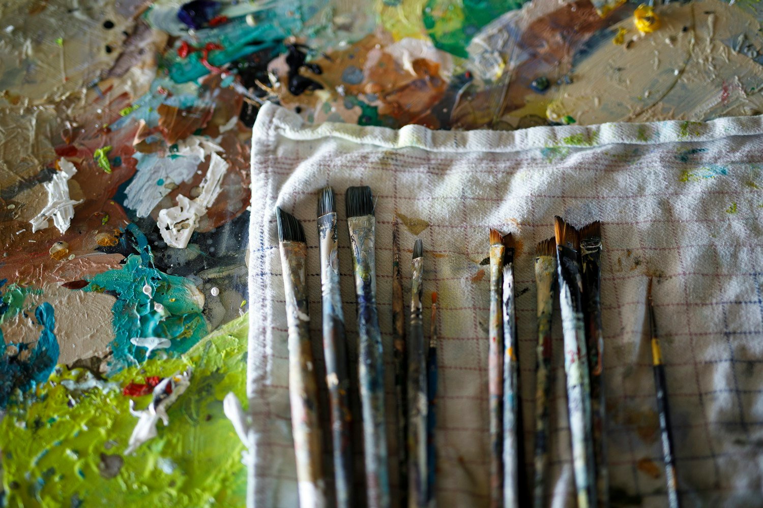 Original photograph of used paintbrushes laid on a cloth beside a painter’s palette